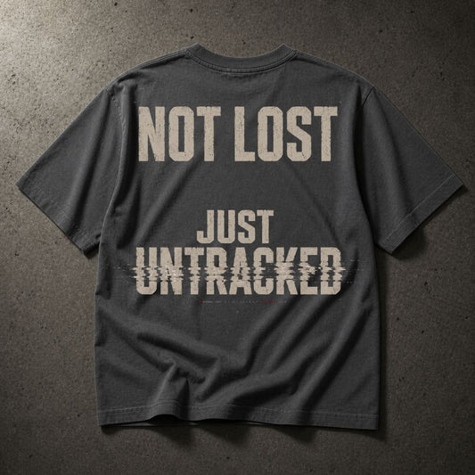 A dark charcoal gray t-shirt laid flat on a textured gray concrete surface, featuring the distressed beige text 'NOT LOST JUST UNTRACKED' printed on the back.