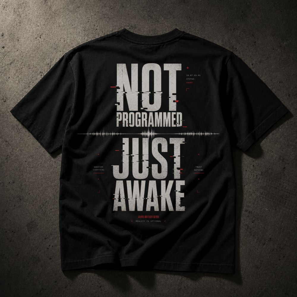 A black short-sleeve t-shirt shown from the back, featuring a large distressed white graphic that reads 'NOT PROGRAMMED JUST AWAKE' with small red and white glitch-style text details, laid flat on a dark textured background.