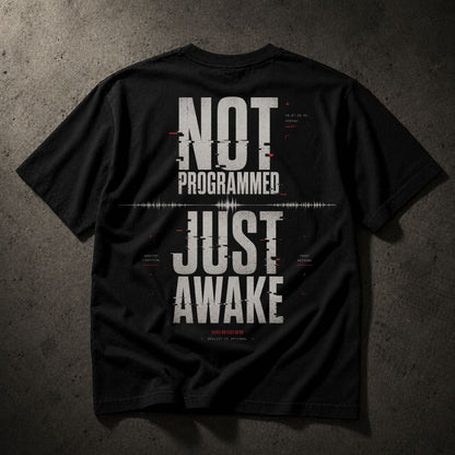 A black short-sleeve t-shirt shown from the back, featuring a large distressed white graphic that reads 'NOT PROGRAMMED JUST AWAKE' with small red and white glitch-style text details, laid flat on a dark textured background.
