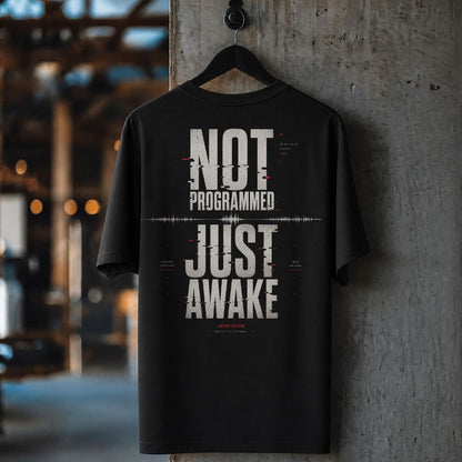 A black short-sleeve t-shirt hanging on a black hanger against a textured concrete pillar in an industrial loft setting. The shirt features a large white graphic text print reading 'NOT PROGRAMMED JUST AWAKE' with a glitch effect and small red accents.