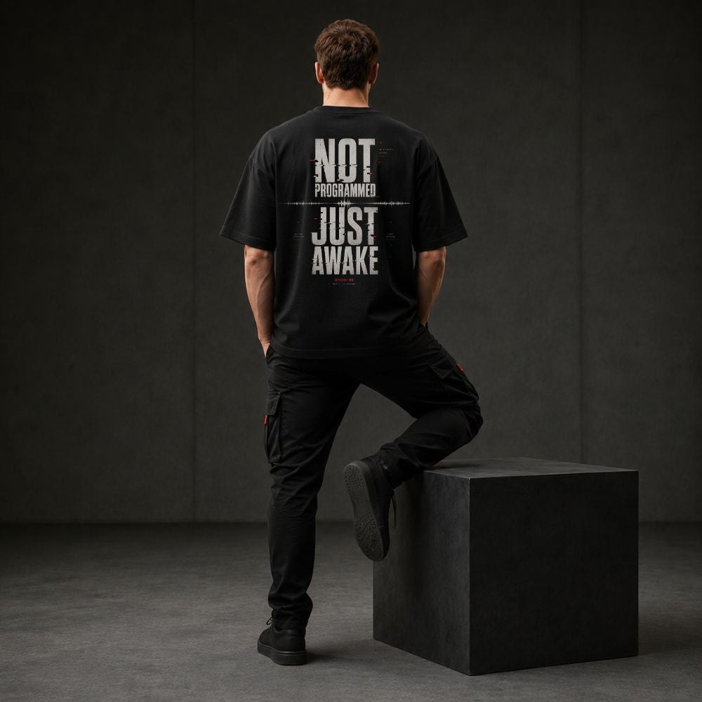 A man stands with his back to the camera wearing a black graphic t-shirt with the text 'NOT PROGRAMMED JUST AWAKE' printed in distressed white letters, paired with black cargo pants and standing on a black block in a dark studio setting.