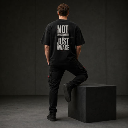 A man stands with his back to the camera wearing a black graphic t-shirt with the text 'NOT PROGRAMMED JUST AWAKE' printed in distressed white letters, paired with black cargo pants and standing on a black block in a dark studio setting.