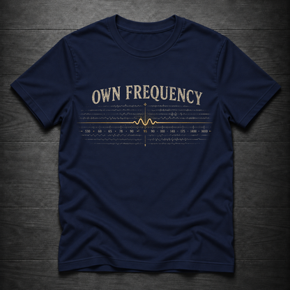 A navy blue short-sleeve t-shirt with a vintage-style cream and gold graphic on the chest reading 'OWN FREQUENCY' above a waveform and frequency graph, laid flat on a dark wood background.