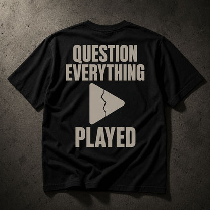 A black short-sleeve t-shirt laid flat on a dark textured concrete surface, featuring the phrase 'QUESTION EVERYTHING PLAYED' in a distressed beige font with a broken play button icon in the center.