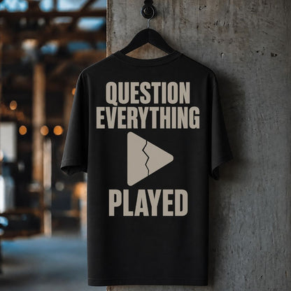A black short-sleeve T-shirt hanging on a black hanger against a textured concrete pillar in an industrial interior. The shirt features a bold graphic print in a warm beige tone reading 'QUESTION EVERYTHING PLAYED' with a broken play triangle symbol.