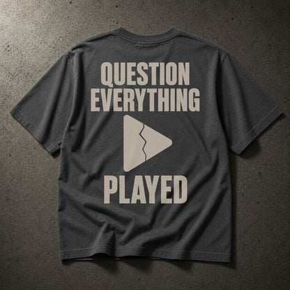 A dark gray t-shirt laid flat on a textured gray surface, showing the back with a light beige graphic that reads 'QUESTION EVERYTHING PLAYED' and a broken play triangle.