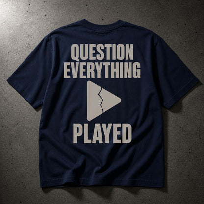 A navy blue short-sleeve t-shirt with a graphic print on the back featuring the text 'QUESTION EVERYTHING PLAYED' in a light beige/gray font and a broken play button icon, laid flat on a dark textured concrete surface.