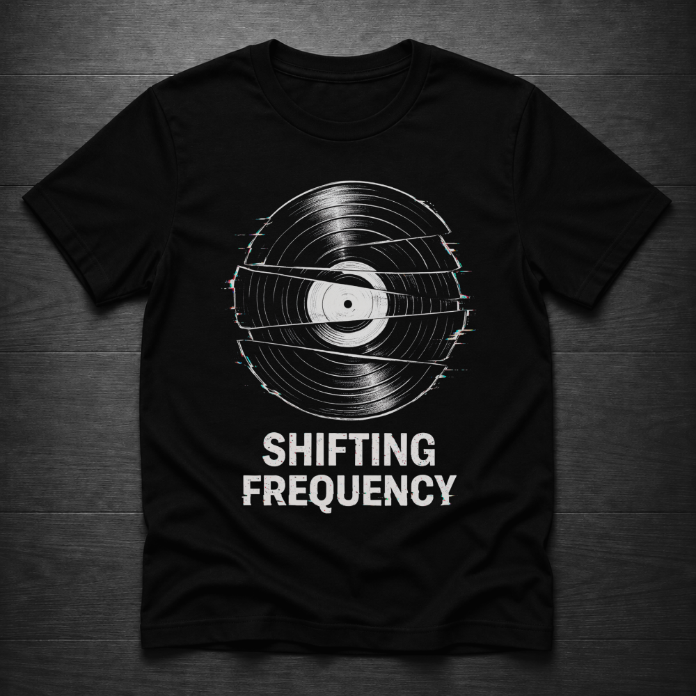 A black short-sleeve t-shirt with a vinyl record graphic and the text 'SHIFTING FREQUENCY' printed in white, laid flat on a dark wood background.