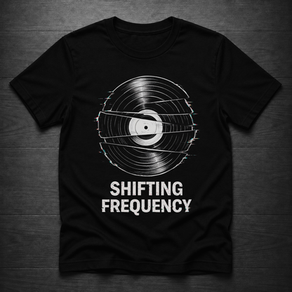 A black short-sleeve t-shirt with a vinyl record graphic and the text 'SHIFTING FREQUENCY' printed in white, laid flat on a dark wood background.