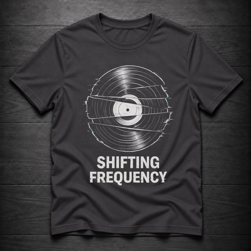A dark charcoal gray t-shirt with a graphic of a vinyl record distorted by horizontal static lines, with the text 'SHIFTING FREQUENCY' printed below in white. The t-shirt is laid flat on a dark wood plank background.