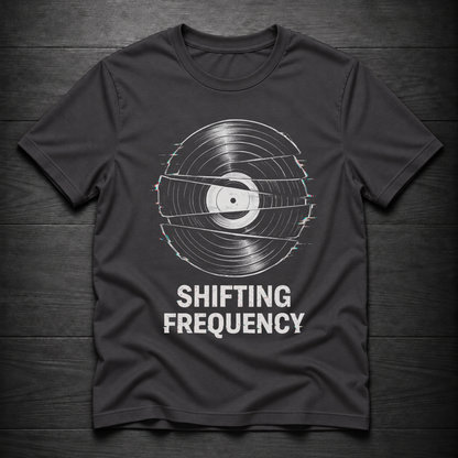 A dark charcoal gray t-shirt with a graphic of a vinyl record distorted by horizontal static lines, with the text 'SHIFTING FREQUENCY' printed below in white. The t-shirt is laid flat on a dark wood plank background.