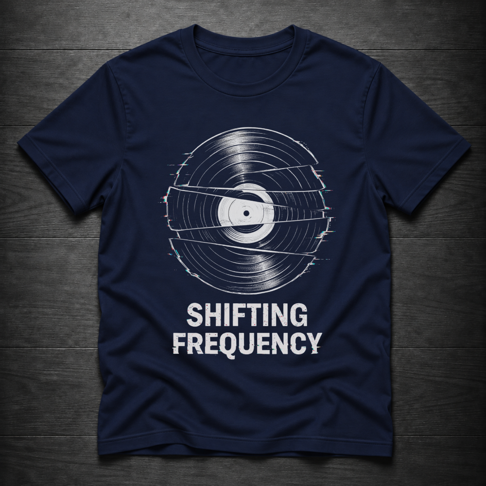 A navy blue short-sleeve t-shirt with a white graphic of a vinyl record with glitch effects and the text 'SHIFTING FREQUENCY' printed below it, laid flat on a dark wood background.