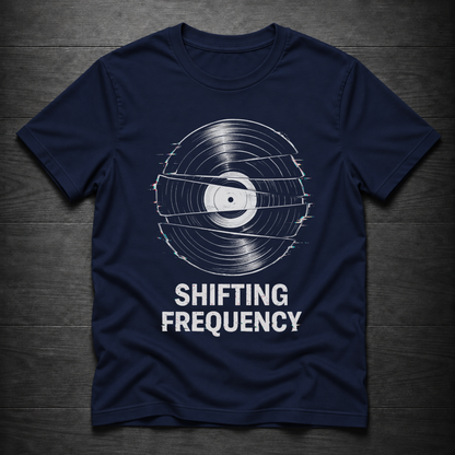 A navy blue short-sleeve t-shirt with a white graphic of a vinyl record with glitch effects and the text 'SHIFTING FREQUENCY' printed below it, laid flat on a dark wood background.