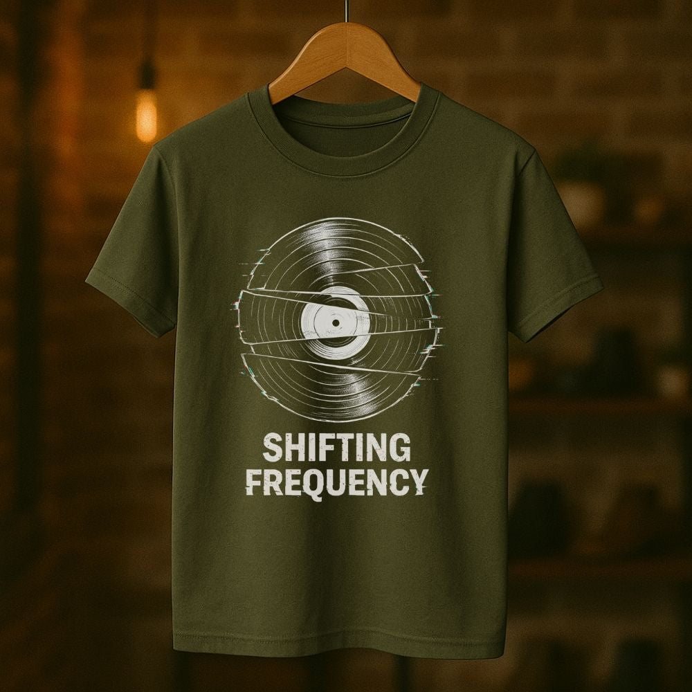 Green t-shirt with vinyl record design and 'Shifting Frequency' text on a wooden hanger against a blurred background.