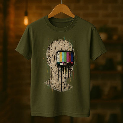 An olive green t-shirt hanging on a wooden hanger against a warm, blurred brick wall background. The t-shirt features a graphic print of a distressed, paint-splattered human head with a television test pattern screen for the eyes.
