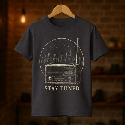 Dark gray t-shirt with a vintage radio graphic and 'Stay Tuned' text on a wooden hanger against a brick wall background.