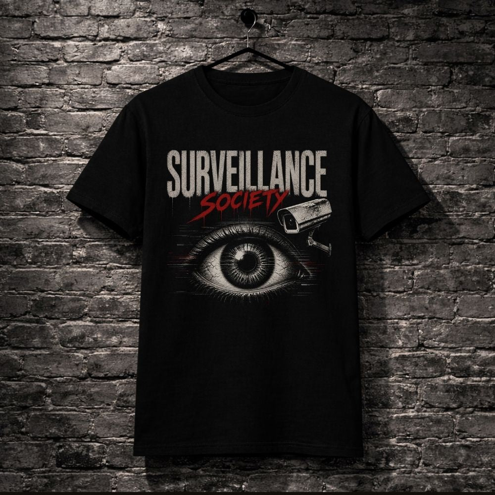 A black short-sleeve graphic t-shirt hanging on a black hanger against a dark gray brick wall. The shirt features a graphic print with the words 'SURVEILLANCE SOCIETY' in white and red, an eye illustration, and a security camera icon.