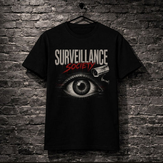A black short-sleeve graphic t-shirt hanging on a black hanger against a dark gray brick wall. The shirt features a graphic print with the words 'SURVEILLANCE SOCIETY' in white and red, an eye illustration, and a security camera icon.