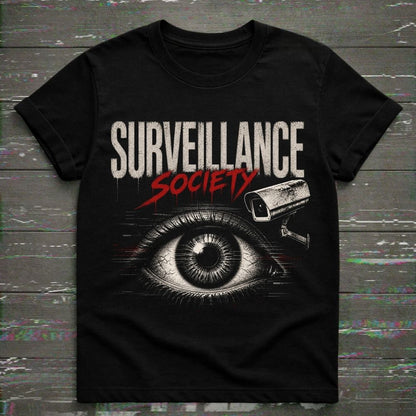 A black short-sleeve graphic t-shirt featuring the words 'SURVEILLANCE SOCIETY' in distressed white and red lettering, with a large illustrated eye and security camera graphic below, displayed flat against a dark wood plank background.