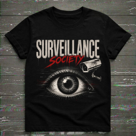A black short-sleeve graphic t-shirt featuring the words 'SURVEILLANCE SOCIETY' in distressed white and red lettering, with a large illustrated eye and security camera graphic below, displayed flat against a dark wood plank background.
