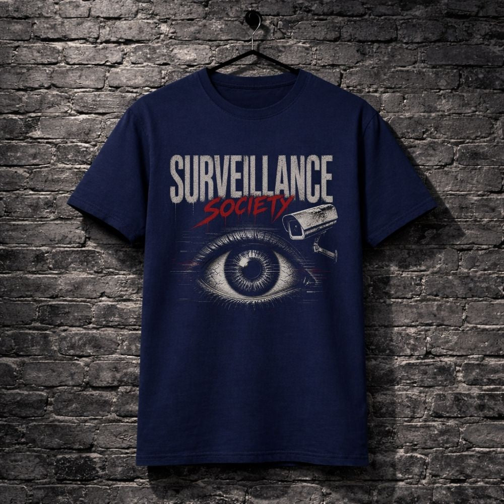 A navy blue graphic t-shirt hanging on a black hanger against a dark gray brick wall. The shirt features a graphic print with the text 'SURVEILLANCE SOCIETY' in white and red, an eye illustration, and a security camera icon.