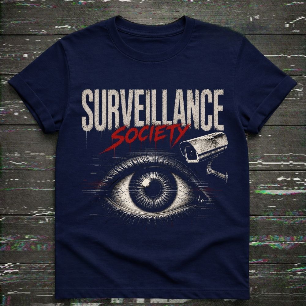 A navy blue short-sleeve t-shirt with a graphic print reading 'SURVEILLANCE SOCIETY' in white and red, featuring a large illustrated eye and a security camera above it, laid flat on a dark wood plank background.