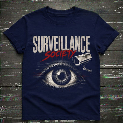 A navy blue short-sleeve t-shirt with a graphic print reading 'SURVEILLANCE SOCIETY' in white and red, featuring a large illustrated eye and a security camera above it, laid flat on a dark wood plank background.
