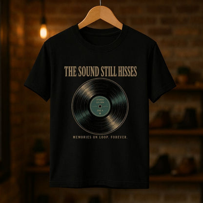 A black short-sleeve graphic T-shirt hanging on a wooden hanger against a warm, blurred brick wall background. The shirt features a vinyl record graphic with the text 'THE SOUND STILL HISSES' above it and 'MEMORIES ON LOOP FOREVER.