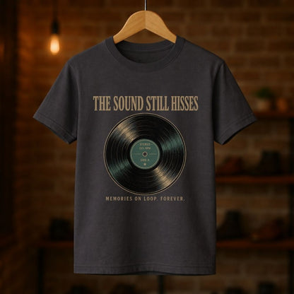 A dark charcoal gray graphic t-shirt hanging on a wooden hanger against a blurred brick wall background. The shirt features a vintage vinyl record graphic in black and green with the text 'THE SOUND STILL HISSES' above and 'MEMORIES ON LOOP FOREVER.