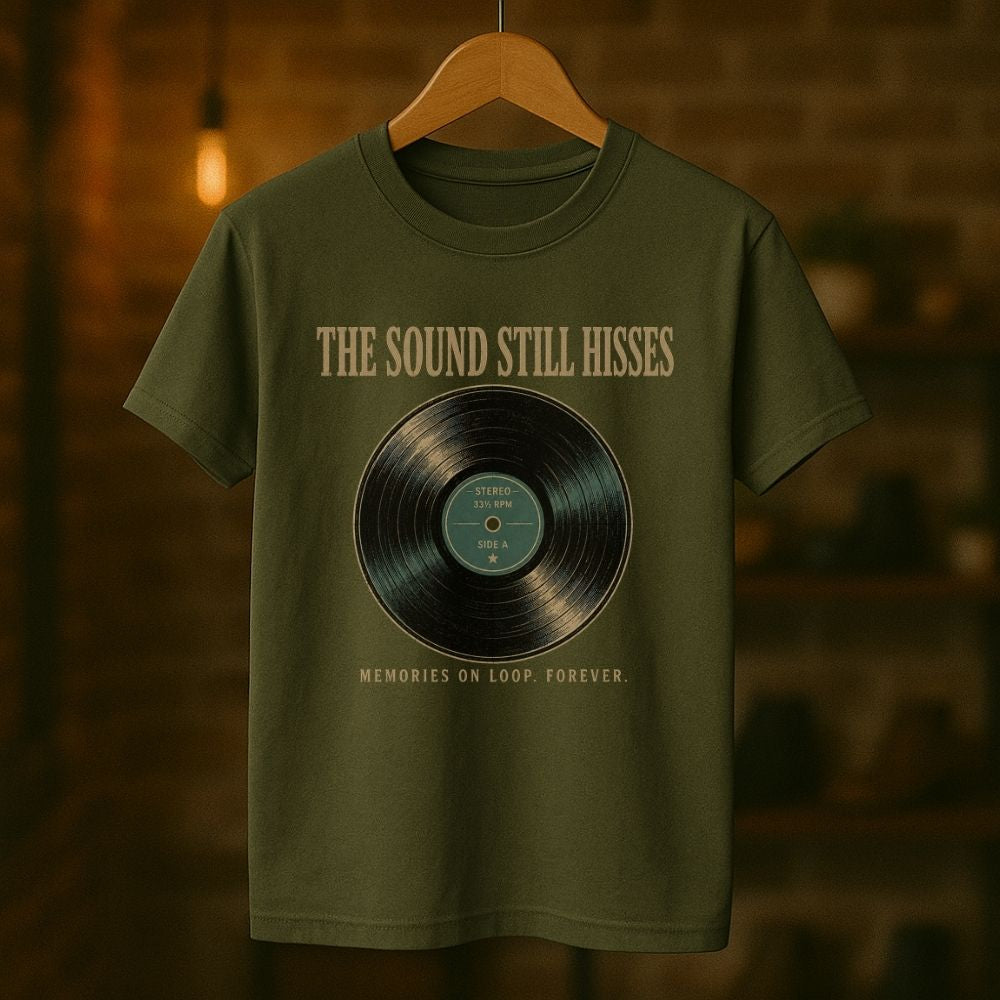 An olive green t-shirt hanging on a wooden hanger against a warm, blurred brick wall background. The shirt features a graphic print of a vinyl record with the text 'THE SOUND STILL HISSES' above it and 'MEMORIES ON LOOP FOREVER.