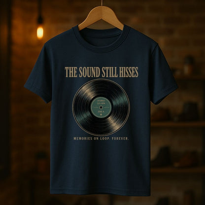 A navy blue graphic t-shirt hanging on a wooden hanger against a warm, blurred brick wall background. The shirt features a vintage vinyl record graphic in the center with the text 'THE SOUND STILL HISSES' above it and 'MEMORIES ON LOOP FOREVER.