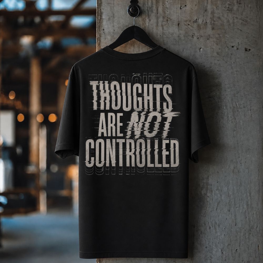 A black short-sleeve graphic T-shirt hanging on a black hanger against a textured concrete pillar in an industrial setting. The shirt features a distressed white typographic print on the back reading 'THOUGHTS ARE NOT CONTROLLED'.