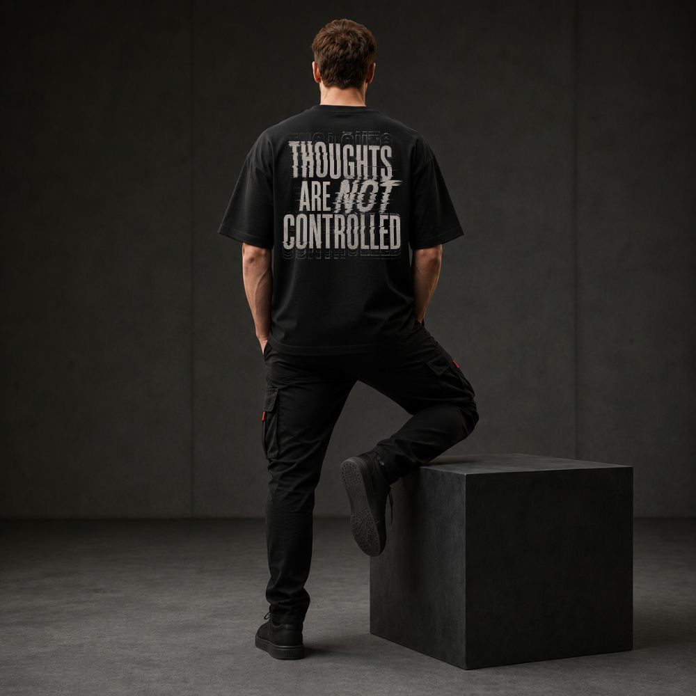 A man stands with his back to the camera wearing a black graphic t-shirt with distressed white text reading 'THOUGHTS ARE NOT CONTROLLED' and black cargo pants, leaning against a black block in a dark studio setting.