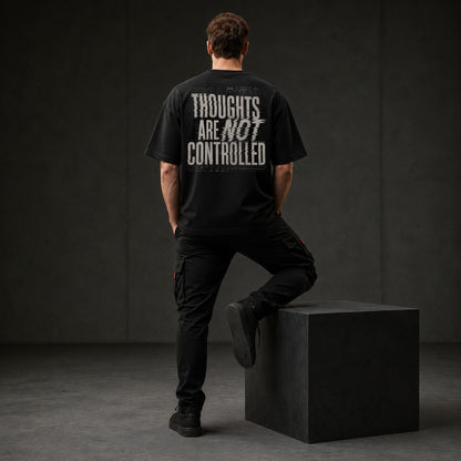 A man stands with his back to the camera wearing a black graphic t-shirt with distressed white text reading 'THOUGHTS ARE NOT CONTROLLED' and black cargo pants, leaning against a black block in a dark studio setting.