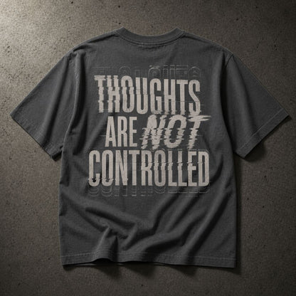 A dark gray t-shirt with a distressed white graphic text reading 'THOUGHTS ARE NOT CONTROLLED' laid flat on a textured gray concrete background.