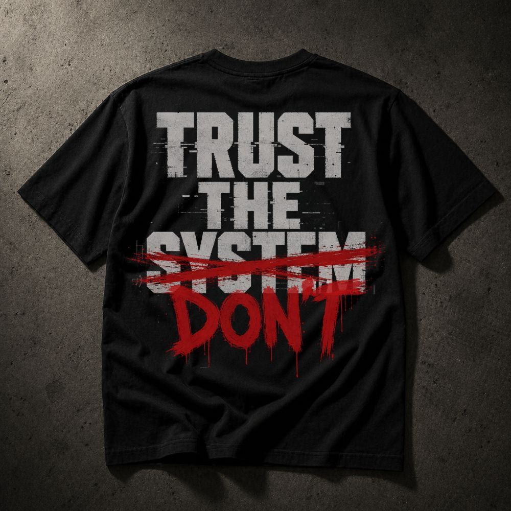 A black short-sleeve t-shirt laid flat on a textured dark gray background, featuring a distressed graphic on the back that reads 'TRUST THE SYSTEM' in white block letters, crossed out with a red brush stroke, and 'DON'T' in red dripping paint below.