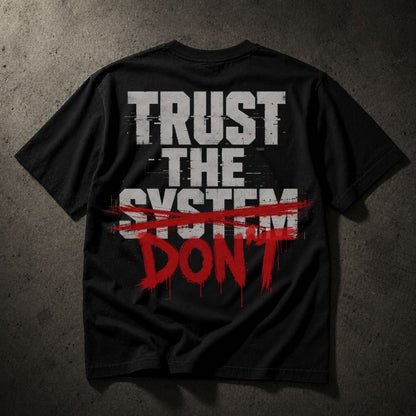A black short-sleeve t-shirt laid flat on a textured dark gray background, featuring a distressed graphic on the back that reads 'TRUST THE SYSTEM' in white block letters, crossed out with a red brush stroke, and 'DON'T' in red dripping paint below.