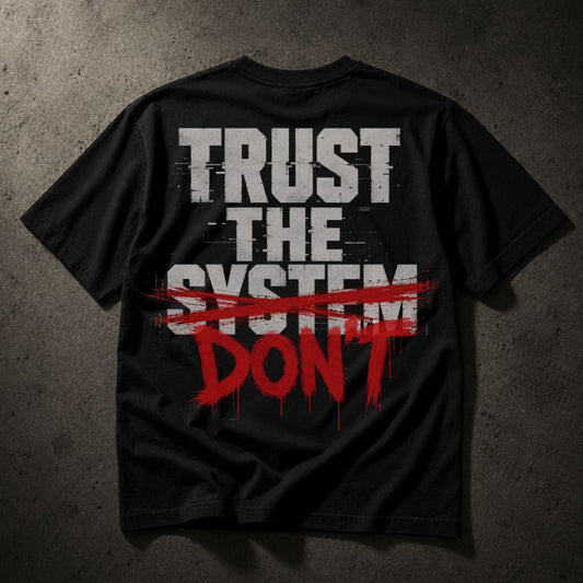 A black short-sleeve t-shirt laid flat on a textured dark gray background, featuring a distressed graphic on the back that reads 'TRUST THE SYSTEM' in white block letters, crossed out with a red brush stroke, and 'DON'T' in red dripping paint below.