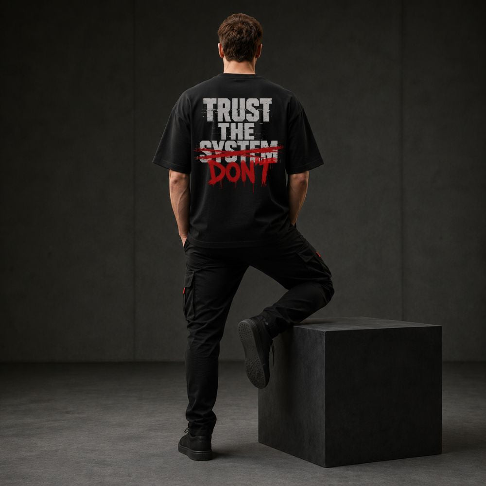 A man stands with his back to the camera wearing a black graphic t-shirt with the text 'TRUST THE SYSTEM DON'T' in white and red, paired with black cargo pants, posing on a black block in a dark studio setting.