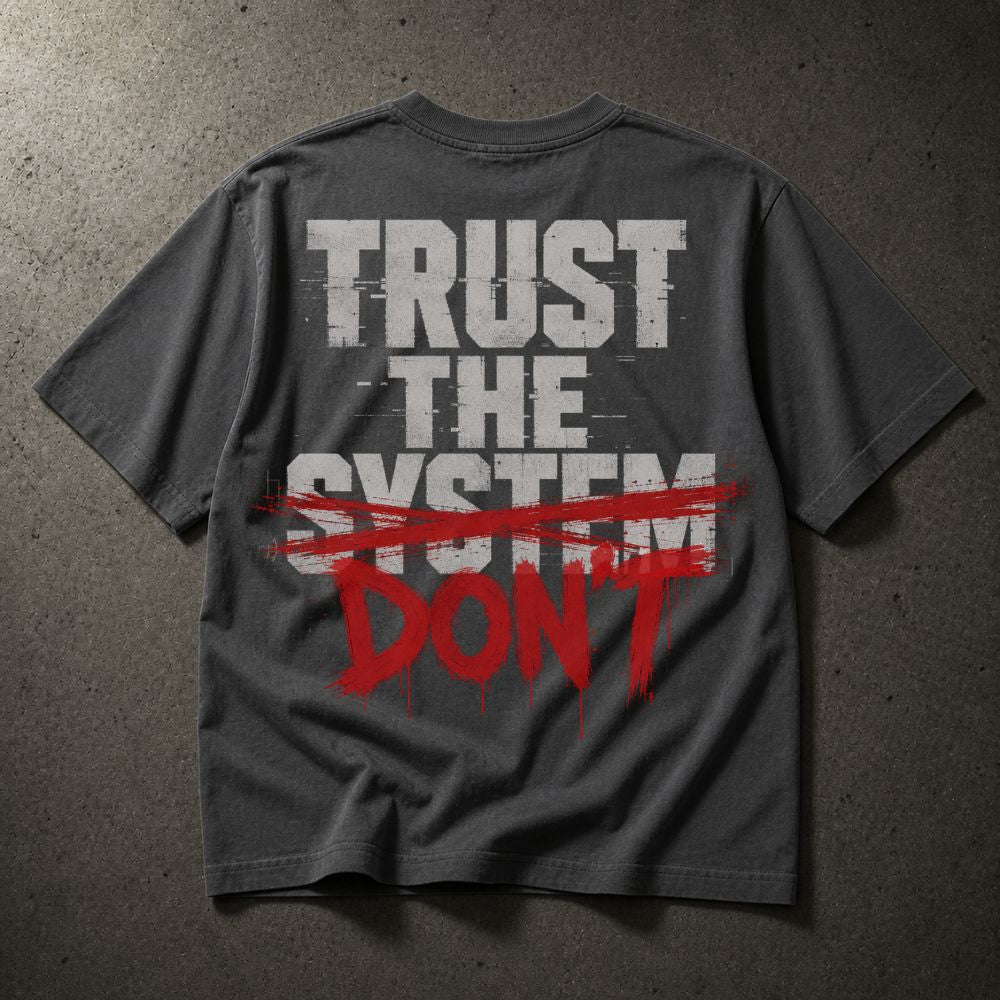 A dark gray t-shirt laid flat on a textured gray background, featuring a graphic print on the back with the words 'TRUST THE SYSTEM' in distressed white letters and 'DON'T' in red paint-splatter style text crossed out with a red brush stroke.