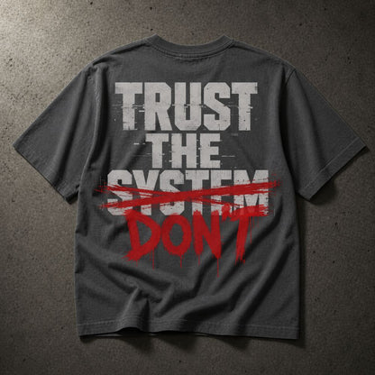 A dark gray t-shirt laid flat on a textured gray background, featuring a graphic print on the back with the words 'TRUST THE SYSTEM' in distressed white letters and 'DON'T' in red paint-splatter style text crossed out with a red brush stroke.