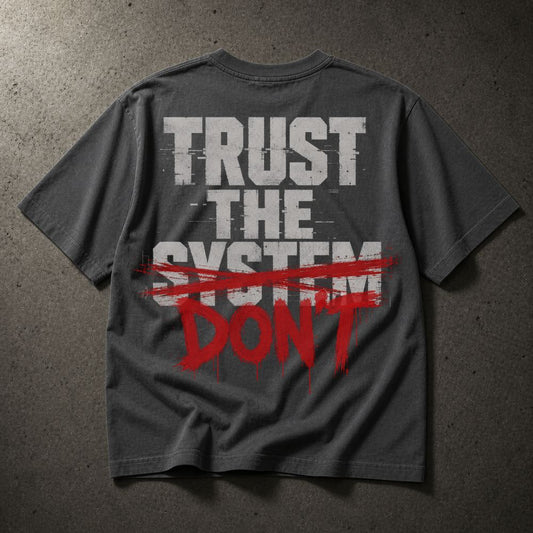 A dark gray t-shirt laid flat on a textured gray background, featuring a graphic print on the back with the words 'TRUST THE SYSTEM' in distressed white letters and 'DON'T' in red paint-splatter style text crossed out with a red brush stroke.