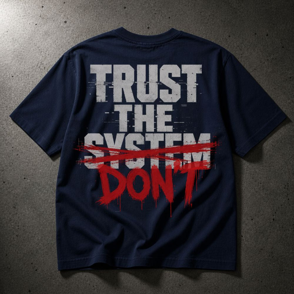 A navy blue t-shirt laid flat on a textured gray concrete background, featuring a graphic print on the back with the words 'TRUST THE SYSTEM' in distressed white letters, crossed out with a red paint stroke, and 'DON'T' written in red dripping paint below.