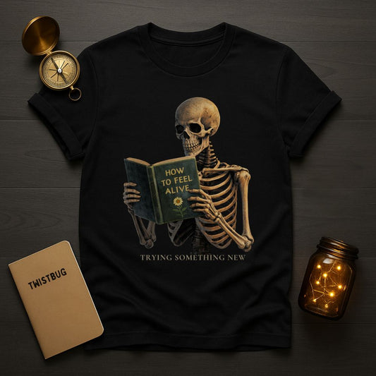 Black graphic T-shirt featuring a skeleton reading a book titled "How to Feel Alive" with the phrase "Trying Something New" on a dark flat-lay background

