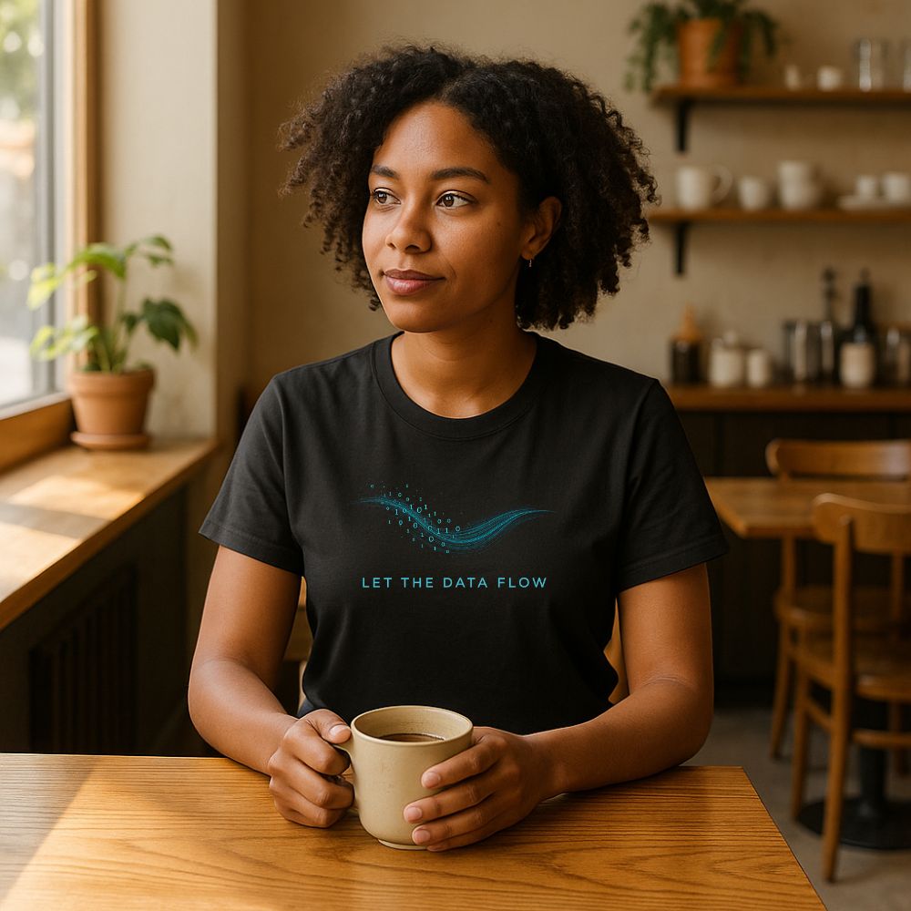 Woman wearing black “Let the Data Flow” T-shirt with blue binary wave design, sitting in cozy café holding coffee.