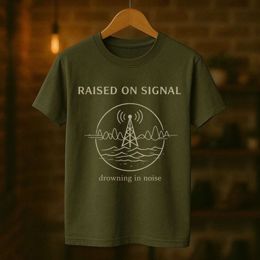 Green t-shirt with 'Raised on Signal' design on a wooden hanger against a blurred background