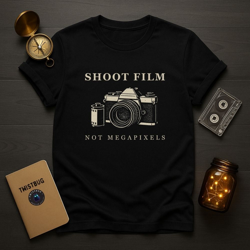 Black Shoot Film Not Megapixels camera design t-shirt laid flat with analog cassette tape and compass accessories.