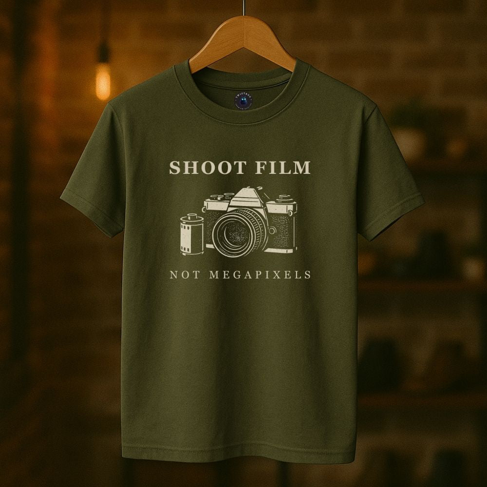 Military green Shoot Film Not Megapixels vintage film camera t-shirt hanging in a rustic shop setting.