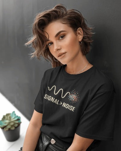 Woman in Twistbug black t-shirt featuring ‘Signal > Noise’ design, sitting by a dark wall with a small plant nearby.