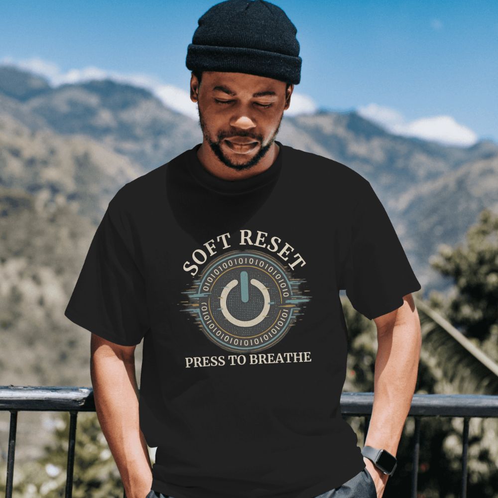 Man wearing black “Soft Reset” T-shirt featuring digital power button design, standing outdoors with mountains in background.